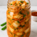 Glass jar filled with homemade keto kimchi made of Napa cabbage, carrots, and chili, placed on a white kitchen counter.